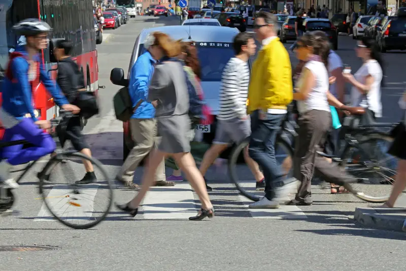 Fotografi på ett övergångsställe. Många människor och cyklister passerar framför biltrafiken. 