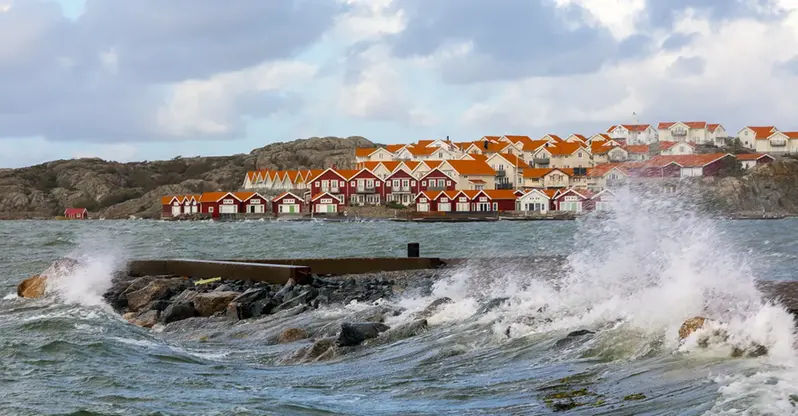 Fotografiet visar ett hav med högt vattenstånd och vågor under en storm. I bakgrunden ett svenskt kustlandskap med många små stugor.