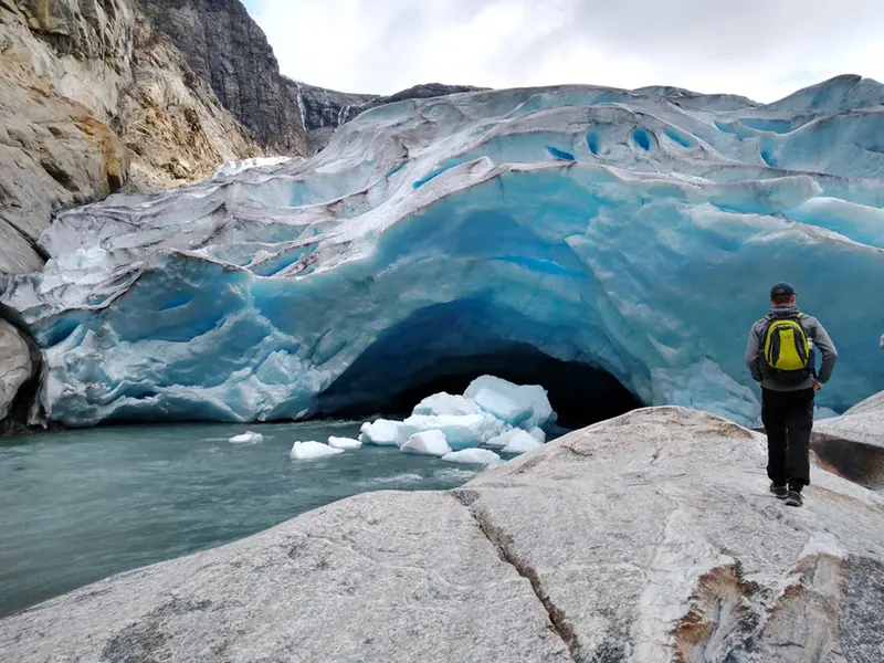 En person står och kollar på en glaciär. En bit av glaciären har fallit ned i en ström av smältvatten.