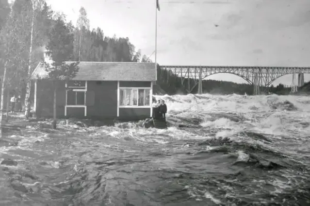 Gråvit bild. Personer i äldre kläder vid forsande älv vid hus.