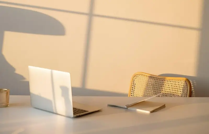 En dator står på ett bord. Solen lyser in och skapar skuggor på väggen.